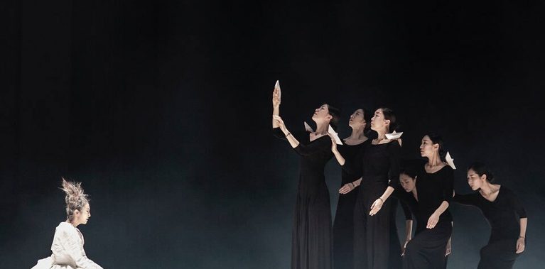 On a dimly lit stage, five dancers in black dresses perform in a triangular formation, gradually lowering in height, each holding a white origami boat. Facing them, a figure dressed in white with an abstract hairstyle sits apart, gazing at the dancers, creating an emotional atmosphere.
