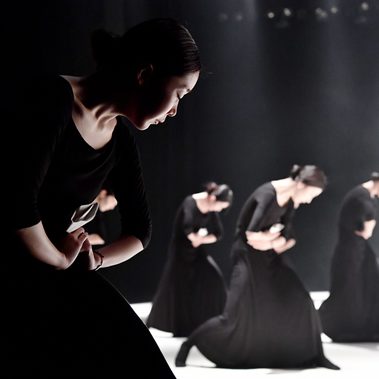 From a perspective view, five dancers in black dresses perform on a dimly lit stage, captured in a synchronised pose with their bodies bent forward and arms laced around their stomachs.