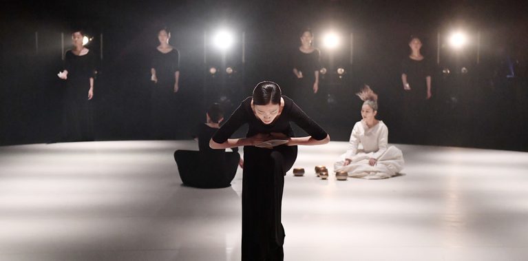 On a dimly lit stage, a performer in black bends forward at centre stage. Behind them, a group of dancers – some standing in line, others seated – creating a calm, introspective atmosphere.