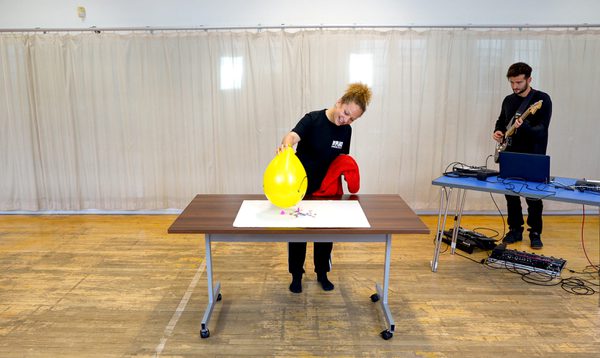 Person in a dance studio holding a yellow balloon upright on a table. Behind them is a person with an electric guitar.