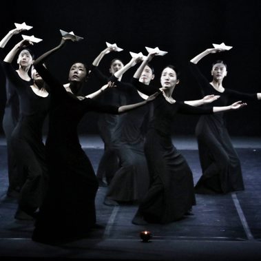Seven dancers in long black dresses perform on a dimly lit stage in unison, each holding a white origami paper boat above their head. They are standing with their legs apart, knees slightly bent, and their left arms extend sideways.