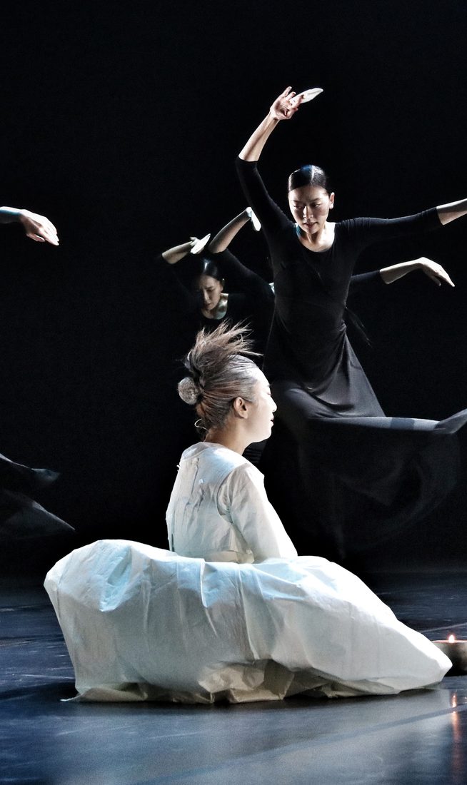 Dancers in long black dresses, each holding a white origami boat, twirl dramatically behind a person in a white costume with a flamboyant hairstyle, seated beside several metallic candles.
