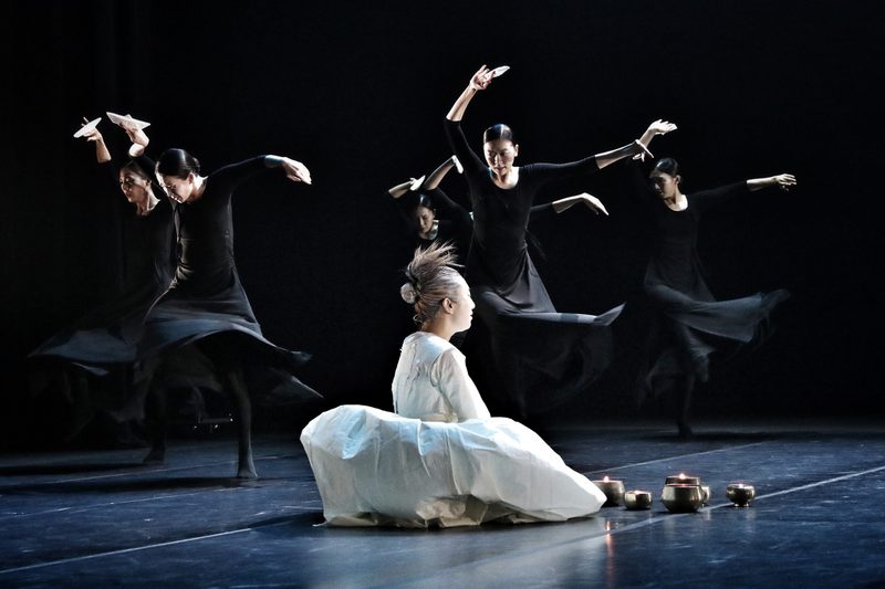 Dancers in long black dresses, each holding a white origami boat, twirl dramatically behind a person in a white costume with a flamboyant hairstyle, seated beside several metallic candles.