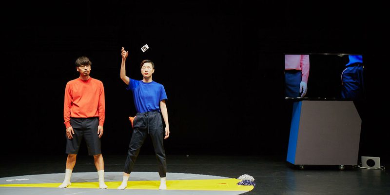 Two dancers stand against a black stage, on a grey circle shot through with yellow. One dancer wears an orange shirt, the other a dark blue shirt. The dancer in dark blue is midway through throwing a small, indistinct object above their head. Their expression is impassive.