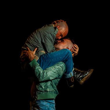 Two male dancers embracing against a black background.