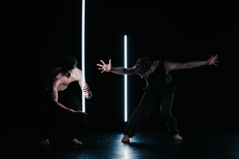 Two male dancers perform a duet with LED light tubes. One crouches near a glowing tube while the other extends his arms wide in a strong stance. Short: Two male dancers perform duet with vertical LED tubes.