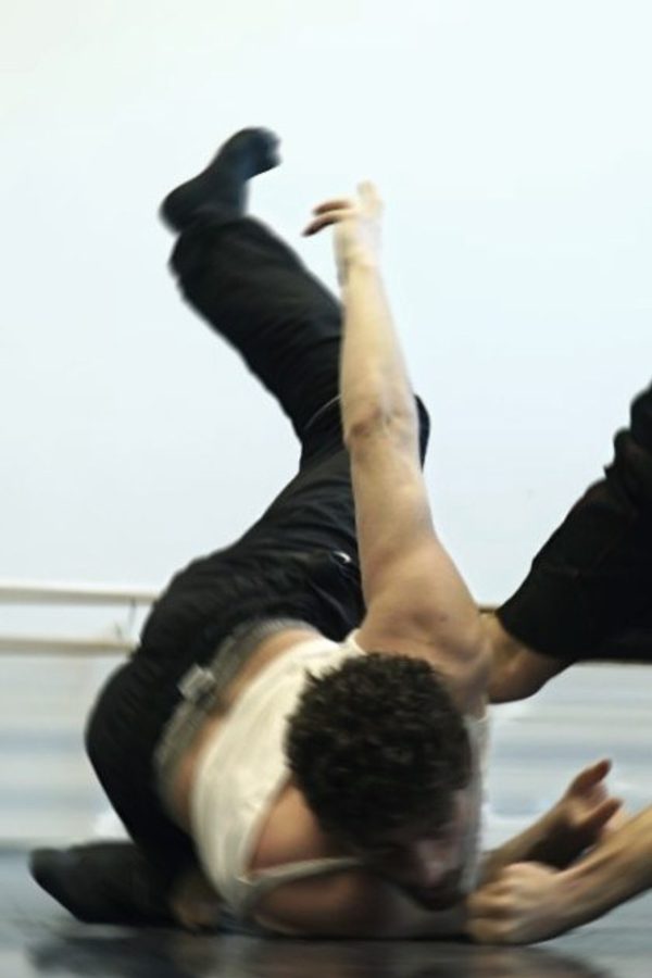 Two dancers in a studio during a floor movement, one low to the ground while the other bends backward over them, limbs extended and motion blurred.
