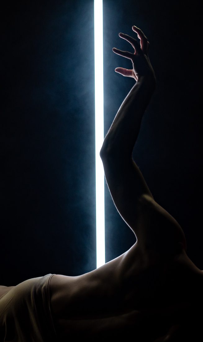 A male dancer lies on the floor, his torso arched back with one arm extended upward. Behind him, a vertical LED light cuts the stage with stark illumination. Short: Male dancer lies back on floor, one arm raised, LED tube behind.