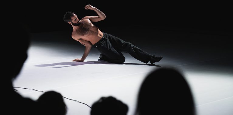 A performer balances low to the floor under a spotlight, supporting themself with one hand while leaning to the side. Their bare torso and dark trousers are illuminated against the dark stage. Audience members are visible in silhouette in the foreground, watching from the edge of the performance space.