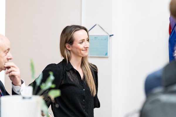 Stephanie stands indoors, smiling and looking to the side. She wears a black, semi-sheer long-sleeved blouse, with other people partially visible around her and a small wall calendar hanging on the wall behind.
