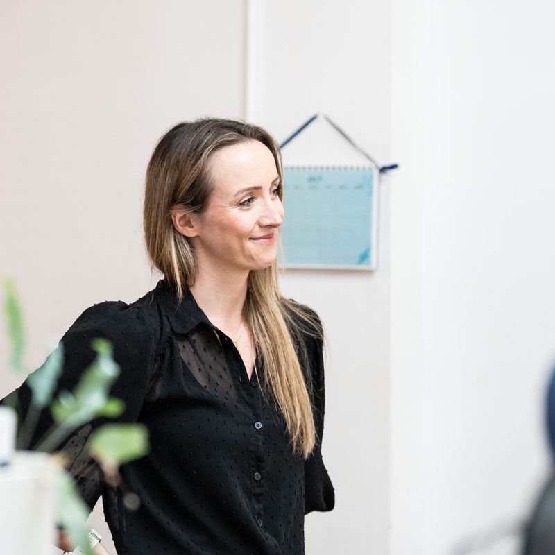 Stephanie stands indoors, smiling and looking to the side. She wears a black, semi-sheer long-sleeved blouse, with other people partially visible around her and a small wall calendar hanging on the wall behind.