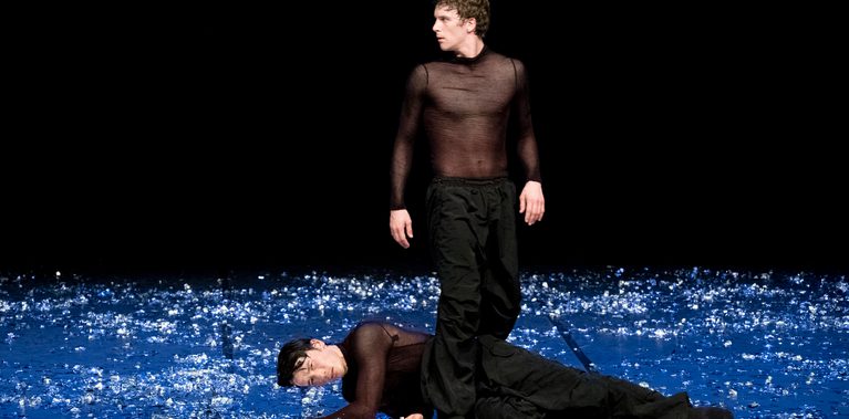 Two male dancers on a stage scattered with reflective fragments. One lies on the ground while the other stands over him, gazing outward into the darkness. Short: Two male dancers; one lies on floor, the other stands over him.