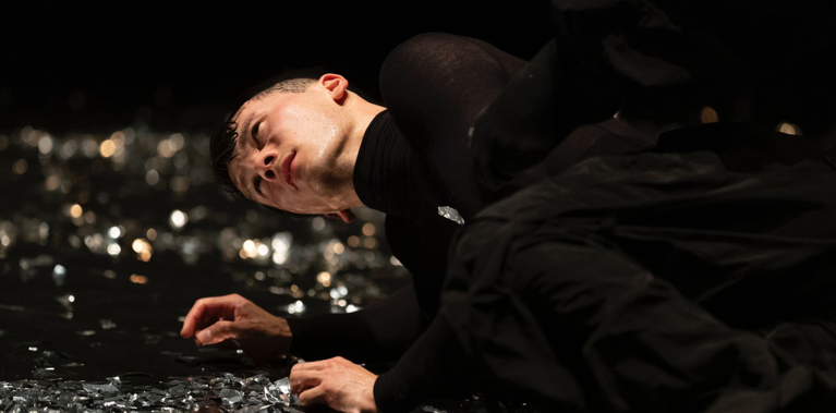 A close-up of a male dancer lying on the stage floor among reflective fragments. His face is turned upward, sweat glistening under the stage lights. Short: Close-up of male dancer lying on stage floor among reflective fragments.