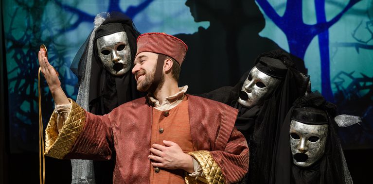 A man in a red and gold costume smiles while holding up a golden ribbon. Behind him, a person dressed in black wears a silver mask and holds up two additional silver masks on either side of the man. A projected forest background with a silhouetted figure appears behind them.