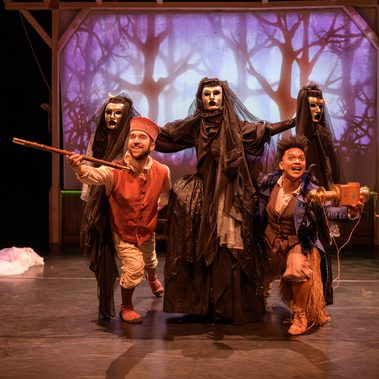 Two performers wearing colourful, folk-style costumes pose at the front—one with a wooden flute, the other with a mechanical prop—while behind them a performer dressed in flowing black and a metallic mask holds two additional veiled, metallic masks raised to create the illusion of three figures. A purple forest backdrop glows behind them with draped white fabric framing the stage.