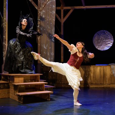 Two performers on stage, one in a black gown on wooden steps reaching forward, the other in a red-and-white dress with one leg lifted.