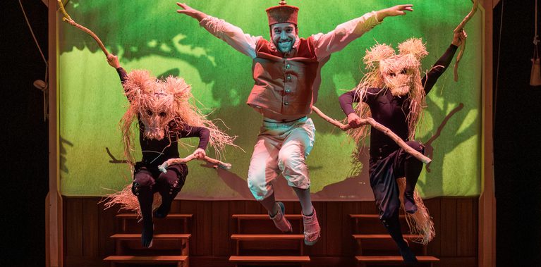 Three performers leap onstage. The central person wears a red vest, white trousers, a small hat, and smiles broadly with arms spread wide. On either side, performers dressed in black wear large straw animal masks and hold long sticks. Behind them a glowing green backdrop featuring a tree-shaped shadow and three sets of small wooden steps.