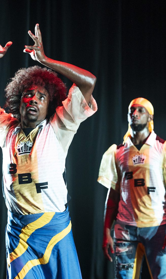 Three performers stand against a dark curtain. The central figure has an afro and wears a blue, yellow, and black wrap over denim, with a white patterned jersey. He looks upward with wide eyes, hands raised in an expressive gesture. Two other men stand behind him in similar jerseys, one wearing a yellow durag and the other with braided hair, both looking on with serious expressions.