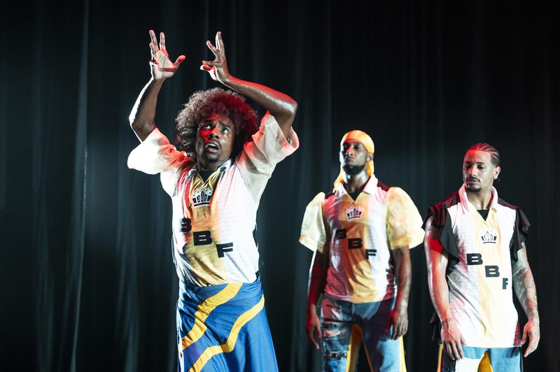 Three performers stand against a dark curtain. The central figure has an afro and wears a blue, yellow, and black wrap over denim, with a white patterned jersey. He looks upward with wide eyes, hands raised in an expressive gesture. Two other men stand behind him in similar jerseys, one wearing a yellow durag and the other with braided hair, both looking on with serious expressions.