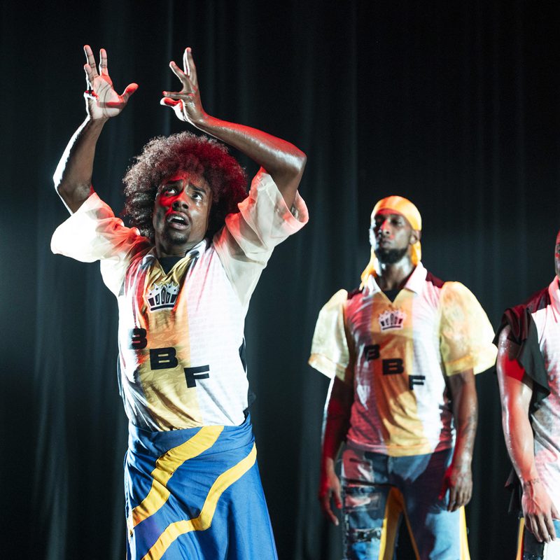 Three performers stand against a dark curtain. The central figure has an afro and wears a blue, yellow, and black wrap over denim, with a white patterned jersey. He looks upward with wide eyes, hands raised in an expressive gesture. Two other men stand behind him in similar jerseys, one wearing a yellow durag and the other with braided hair, both looking on with serious expressions.