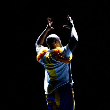 A person in a white and yellow shirt with ruffled orange shoulders and blue and yellow striped pants stands against a black background, arms raised with hands open, illuminated by dramatic top-down lighting.