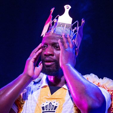 A close-up shot of a performer in a white and yellow jersey wearing a large, ornate crown made of gold-toned afro combs. His face is glistening with sweat under bright blue and purple stage lights as he holds his hands to the sides of his head with a focused, intense expression.