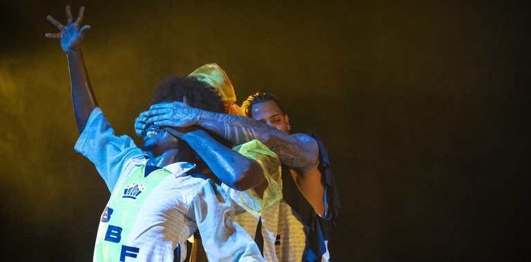 Three performers stand against a black backdrop. The central figure wears a blue, yellow, and white and looks upward with raised hands. Two men stand behind him in matching attire.