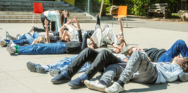 Young teens lying on the ground, gazing and pointing at the sky in front of their school on a bright day.