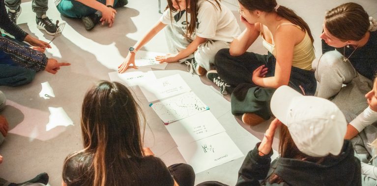 A group of young teens, sitting in a circle on the floor, discussing notes and drawings, with one girl explaining.