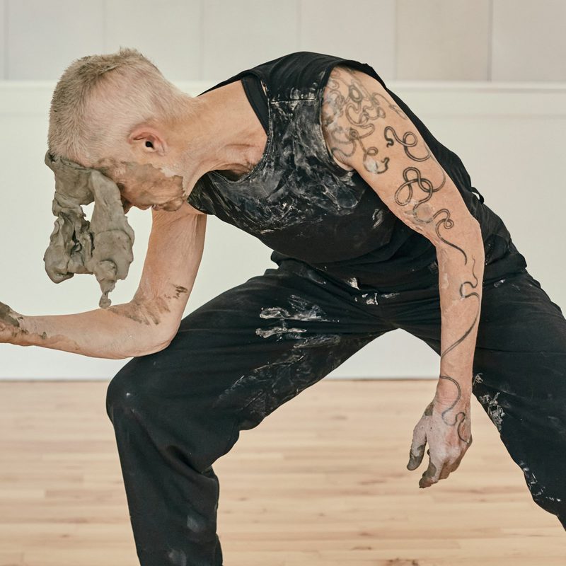 A woman with short hair is wearing a black tank top and trousers has a large chunk of clay on her face and is leaning over with her elbow on her knee.