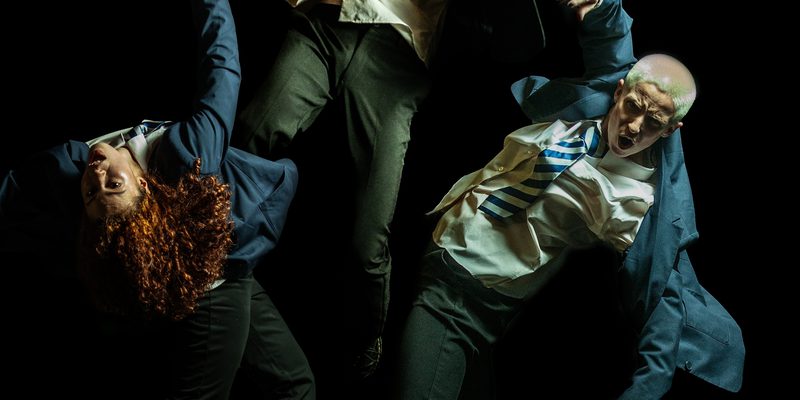 Three dancers in expressive poses mid-air, uniformed in blazers and striped blue ties, their faces strained.