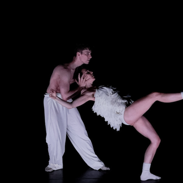 A duet on a bare stage: a male dancer in white trousers supports a female dancer in a textured white costume. She leans back, her leg extended while his hand steadies her head. Short: Male dancer supports female dancer in white costume leaning back.