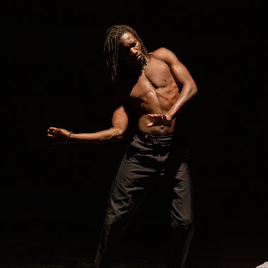 A performer stands shirtless under a spotlight on a dark stage, their body angled to one side with arms extended in a sculptural pose. The lighting highlights the contours of their torso while the surrounding space remains in shadow.