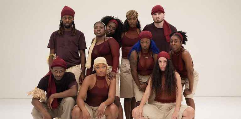 Nine performers pose together for a group portrait in a bright white studio. Arranged in two rows, they face the camera with serious expressions and wear coordinated burgundy and beige costumes.