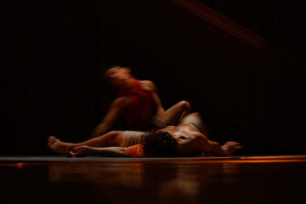A blurred, long-exposure shot captures two dancers in motion on a dark stage. One dancer lies on the floor while the other, wearing red, appears as a streak of movement above them. The warm, low-key lighting emphasizes the fluid and abstract nature of the choreography.