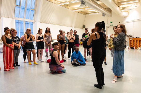 A group of people in a dance studio stand and sit in a semi-circle, attentively listening to two people standing at the front, with Polly on the right of the two as they address the group. The studio has high ceilings, large windows, and an open floor, with participants dressed in various workout and dance attire.