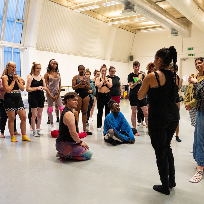 A group of people in a dance studio stand and sit in a semi-circle, attentively listening to two people standing at the front, with Polly on the right of the two as they address the group. The studio has high ceilings, large windows, and an open floor, with participants dressed in various workout and dance attire.