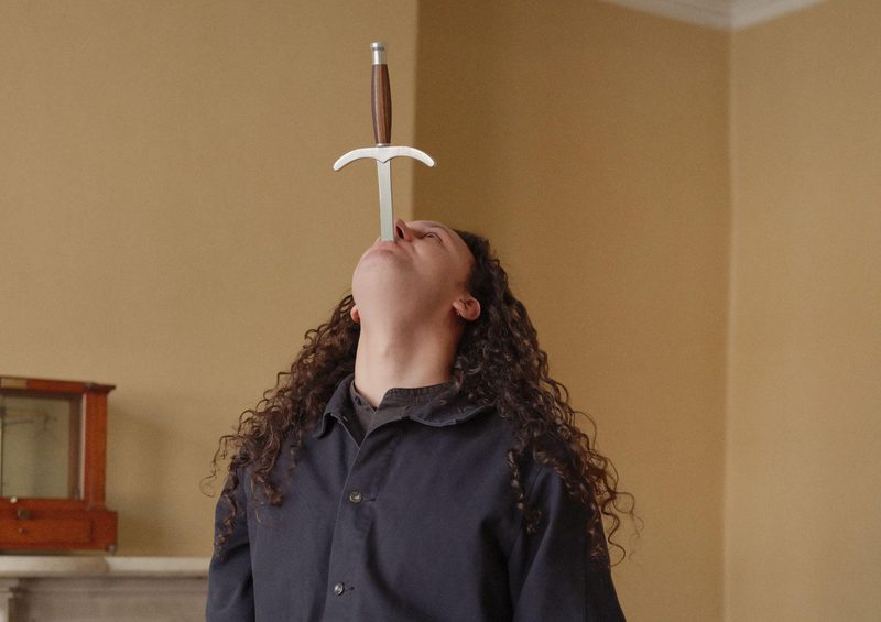 Photo of Tom Cassani in the middle of swallowing a sword, with the handle and upper section of the sword still visible. He is looking upward, has long curly brown hair, and is wearing a black long-sleeved button-up shirt. The image is taken indoors against plain yellow walls.
