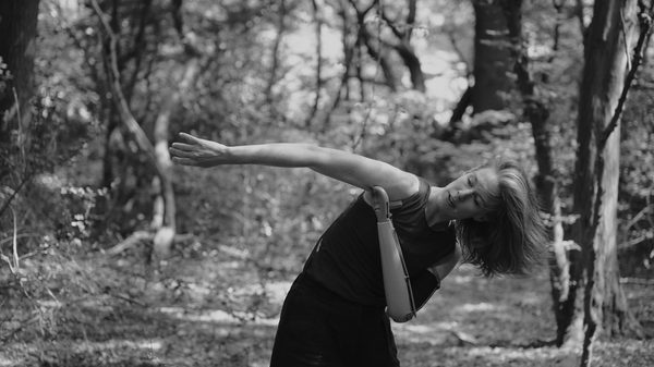 Black & white image of Annie, a tall white person in her late 30s, dancing outside in the forest, with trees behind her. Her torso curves to the left as her right arm reaches out to her right, her left prosthetic arm bending at the elbow to touch her right shoulder. Eyes open and face turned to look towards her arms, shoulder-length hair hanging down to the ground from the momentum of the movement. She's wearing a sleeveless top and dark trousers.