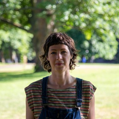 A close-up shot of a white woman in her 30s. She has short medium brown hair with a short fringe and is smiling against a background of trees in a park.