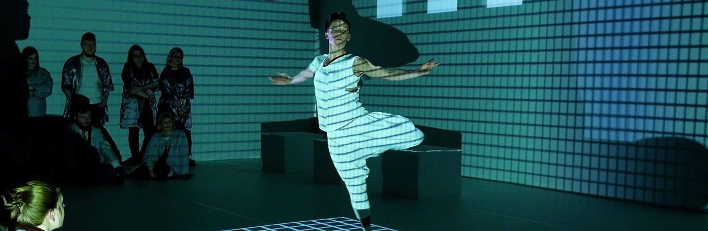 A dancer in a white vest and trousers pirouettes in a dark room, a grid of light projected onto him and the walls behind him. Audience members can be seen around the frame of the image.
