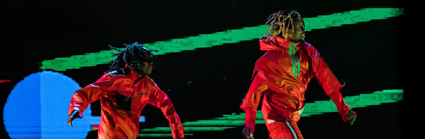 Image of two dancers in the frame. They both are wearing red tracksuits and black trainers. In the background behind them is a blue circle to the left. Two green lines also appear in the background.