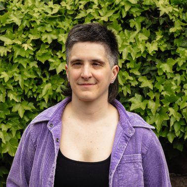 Amelia is wearing a black top with a purple shirt over the top. They are standing against a vibrant green hedge.