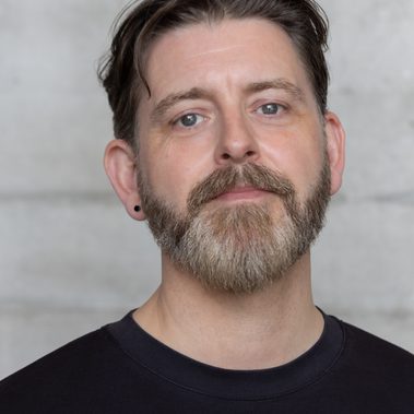 A man wearing a black t-shirt. He is facing the camera head on and only his head and shoulders are in frame.