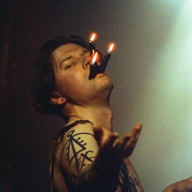 A man is topless and holding three candles in his as his hand reaches up to the sky.