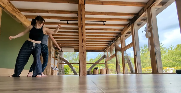 Two barefoot women balance against each other's backs inside a sunlit wooden studio with large windows. Green trees are visible outside, and exposed wooden beams frame the open space.