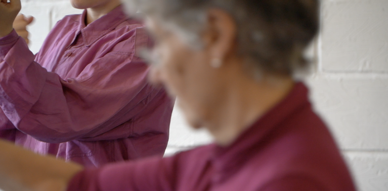 Two people, one blurred in the foreground and the other in focus in the background. Both wear pink clothes. The person in the foreground has grey hair and wears glasses, the person in the background appears younger.