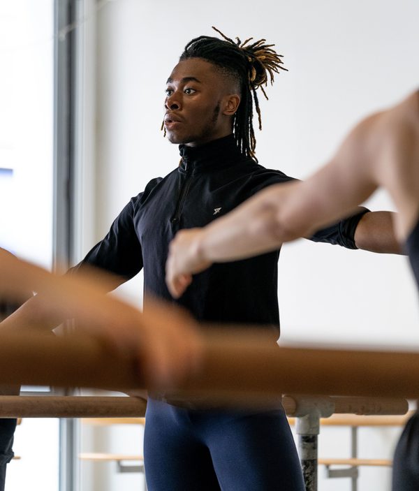 In the background a dancer holds on to a ballet barre with their arm outstretched to the side. They are looking directly forwards with focus and wearing all black. There are other arms in the foreground