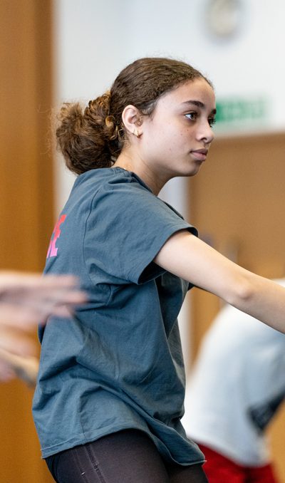 A dancer with brown hair, wearing a grey t-shirt has their arms in a curved position in front of their chest. They are looking forwards towards the mirrors in the studio
