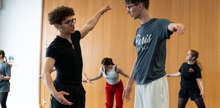 Two young students wearing glasses and dance clothing are working together. They have their arms outstretched to the side and are speaking to each other. In the background there are three other dancers